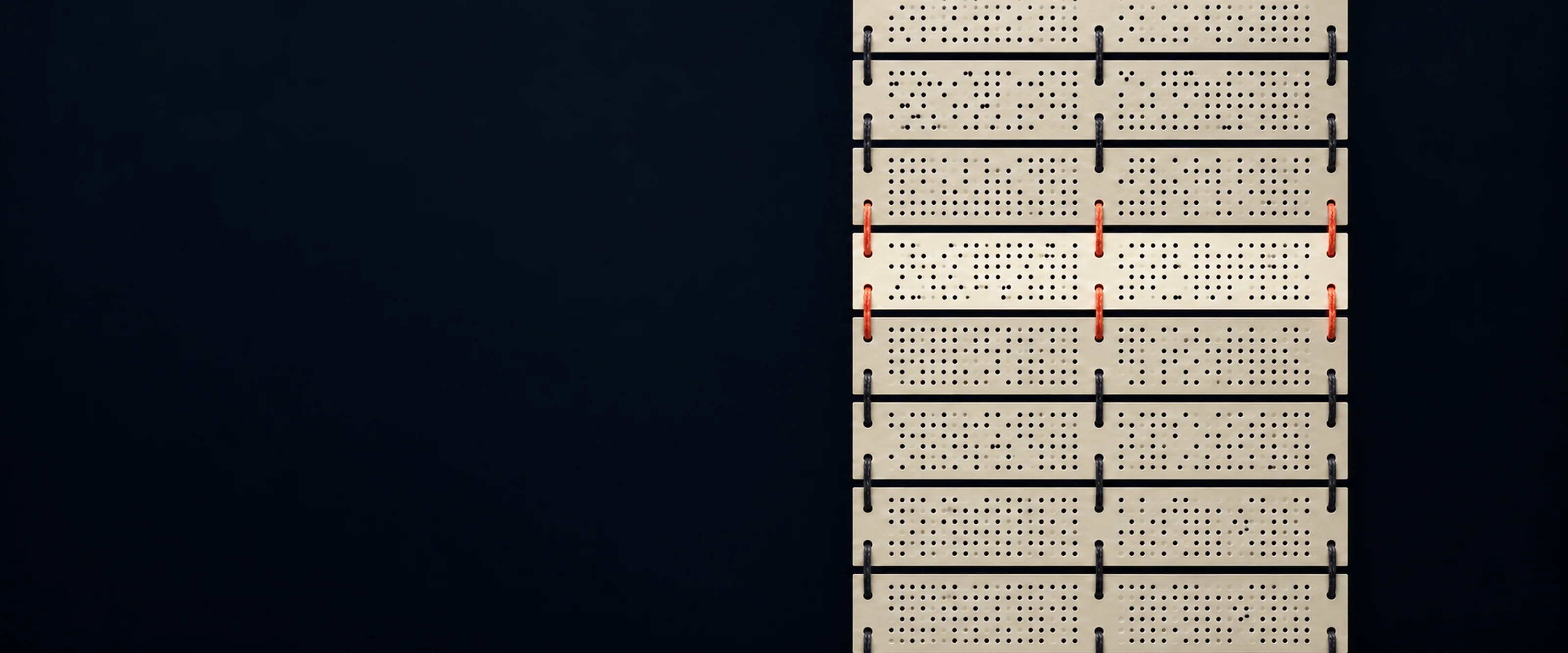 A chain of ivory punched cards laced together with dark thread hangs in the dark. A new card in the chain is laced in with coral cord.