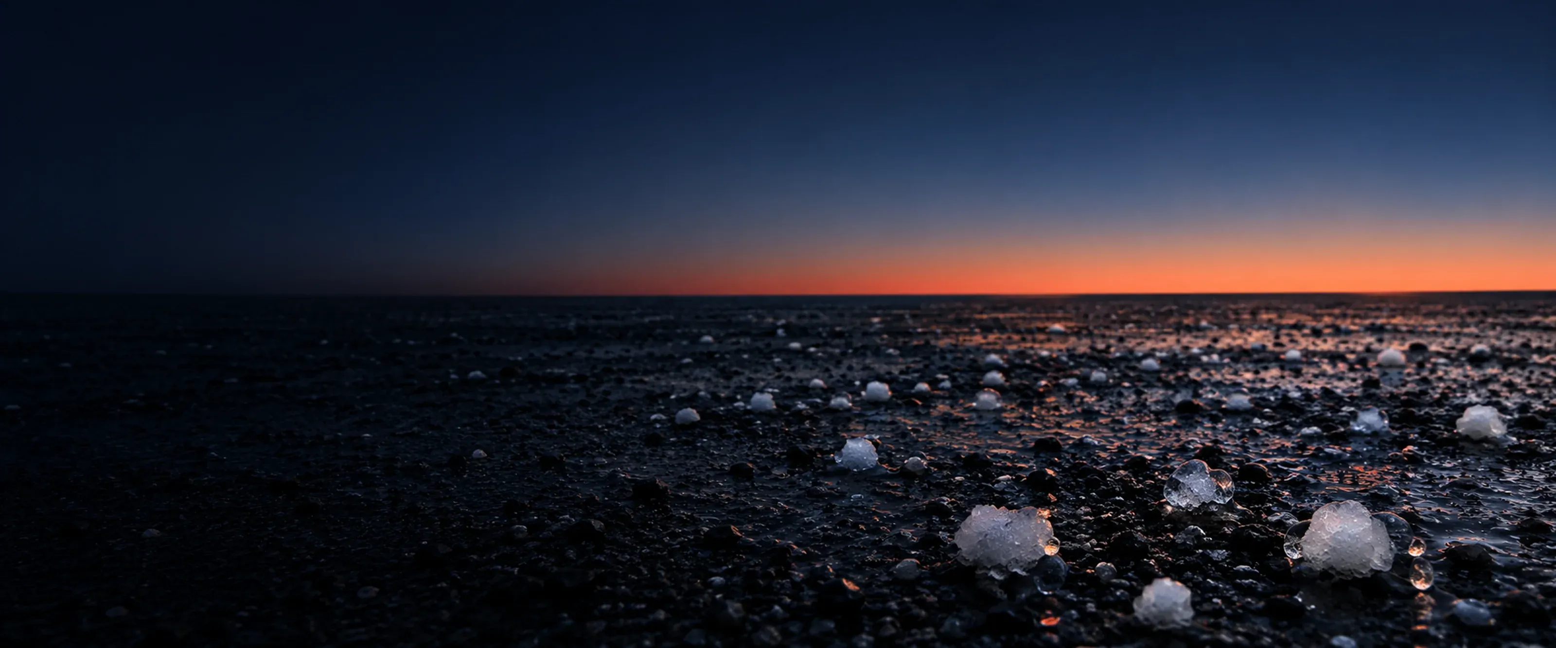 The same dark plain after the storm under a clear evening sky, coral along the horizon. In the foreground the last hailstones melt into beads of water.