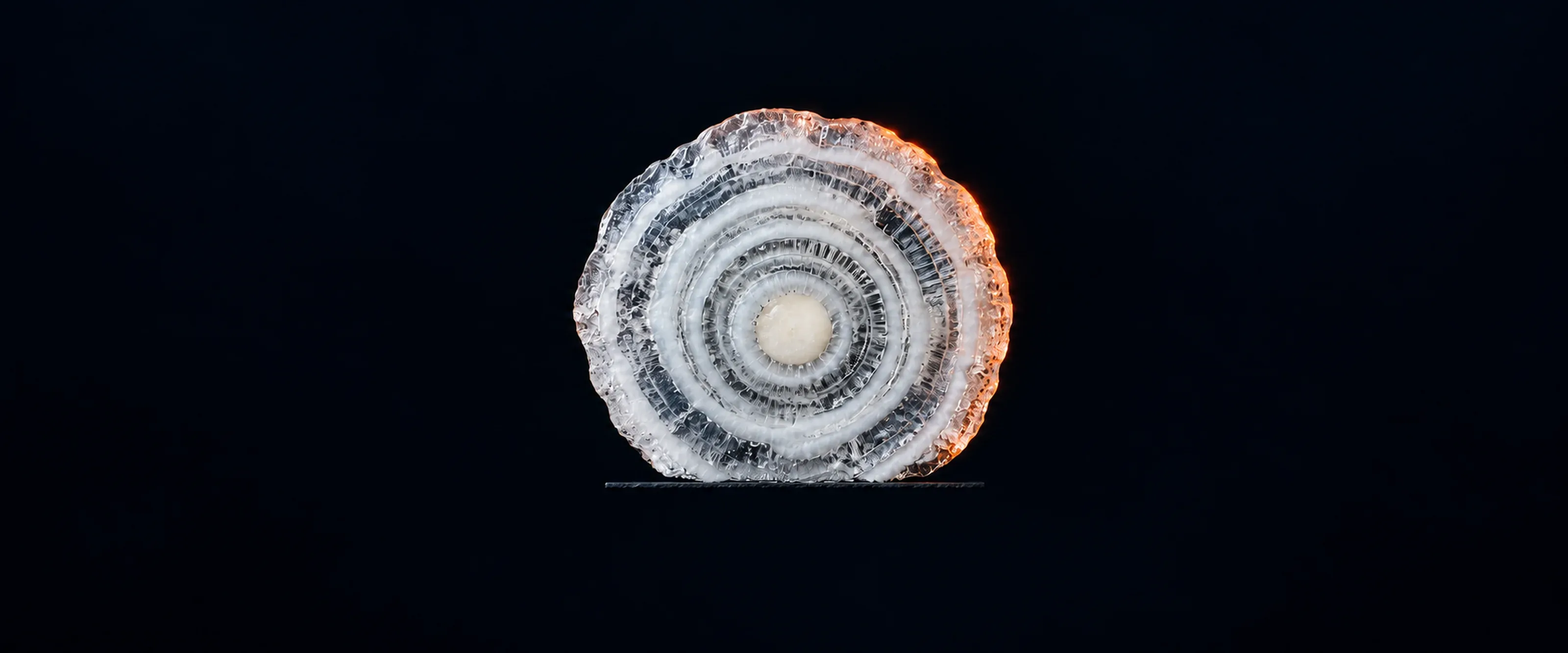 A hailstone cut in half, its cut face showing rings of clear and milky ice around a white core. A thin rim of coral light catches the outermost ring.